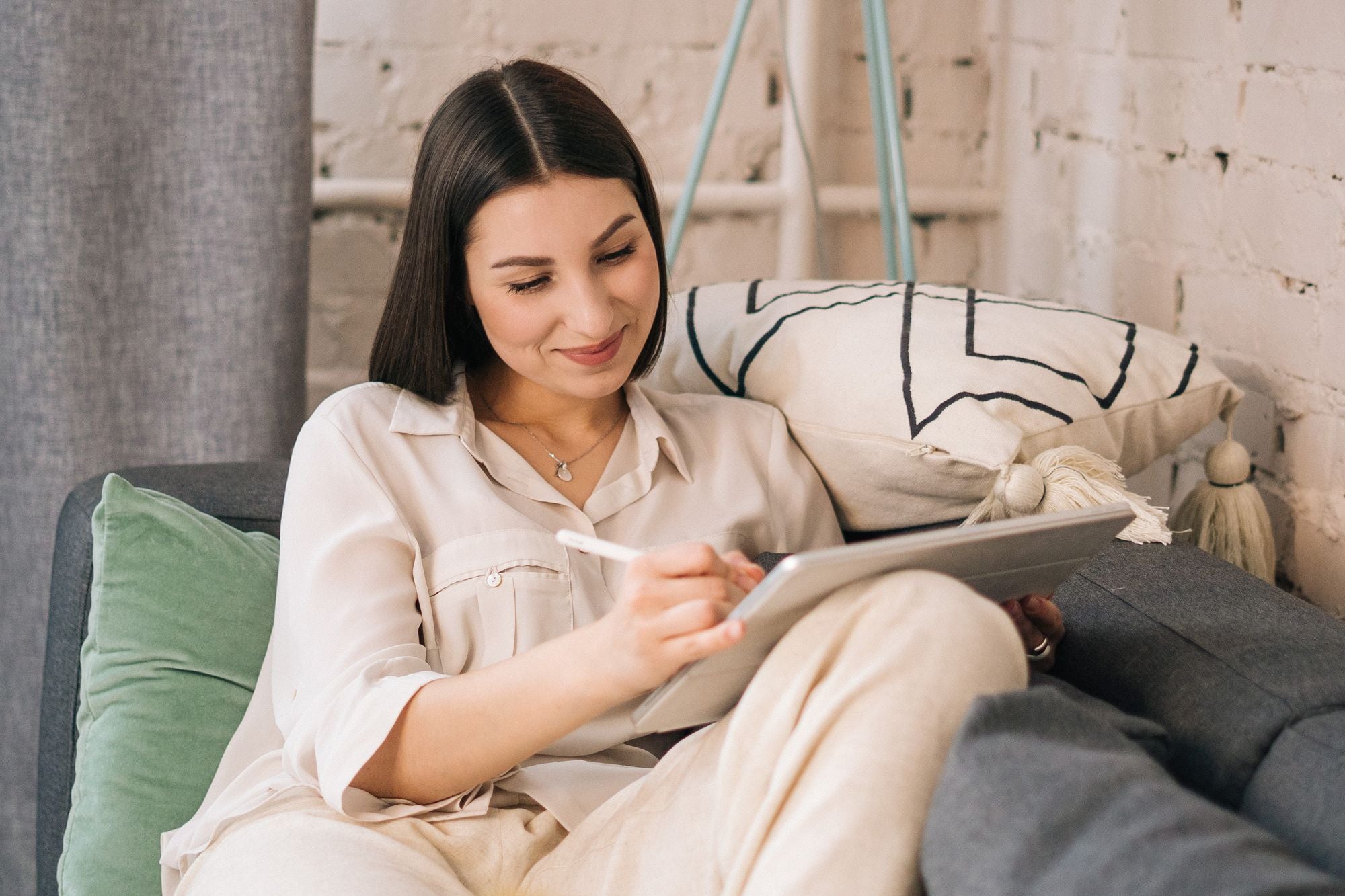 A woman uses a tablet while sitting on a grey sofa.